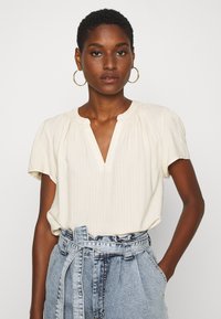 Krämfärgad kortärmad blus med V-ringning och detaljerad fållning vid halsen, i kombination med högmidjade denimbyxor med bälte.