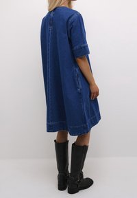 Jeansklänning, lös passform, knälängd, korta ärmar, blå färg, oklippta kanter, sidofickor. Bärs med höga svarta läderstövlar.