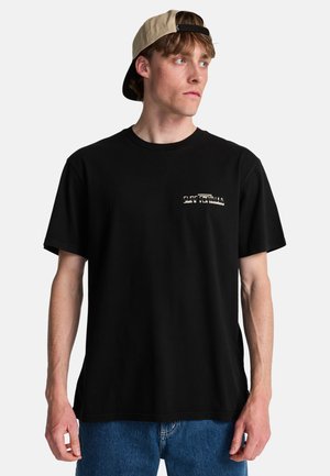 Młody mężczyzna w czarnej koszulce Surf Formula i beżowej czapce z daszkiem założonej do tyłu, patrzący w prawo z neutralnym wyrazem twarzy.