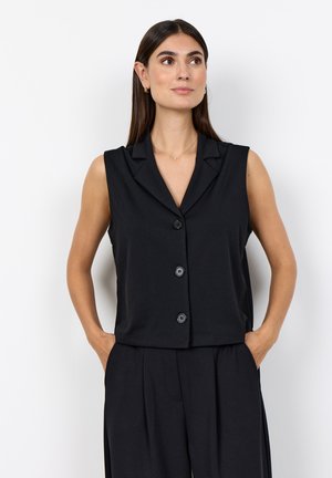 Woman with straight dark hair wears sleeveless black buttoned vest and black pants, standing with hands in pockets against white background.