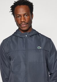 Námornícky vetrovka s zapínaním na zips, kapucňou a malým logom Lacoste. Tkanina má jemnú štruktúru a vertikálne švy.