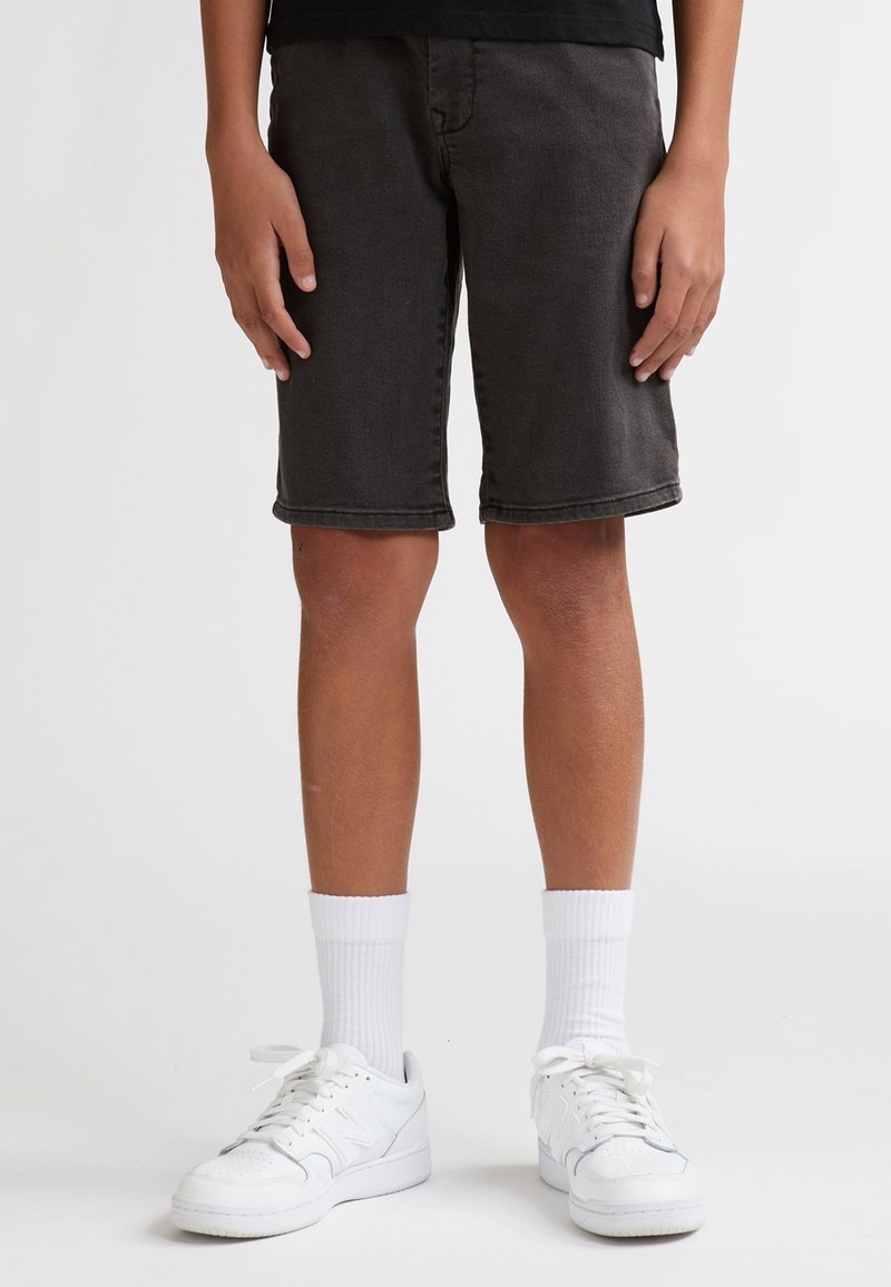Shorts en jean gris foncé avec une coupe classique, longueur au genou, ourlets retroussés, associés à des baskets blanches et des chaussettes blanches mi-mollet.