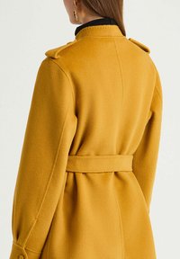 Manteau en laine jaune moutarde, silhouette cintrée, taille ceinturée, épaulettes, texture lisse et détails sur les manches.