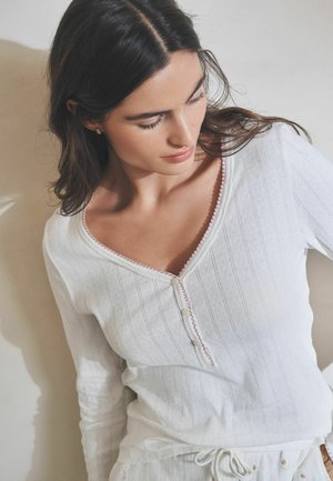 Sötét hajú nő fehér, hosszú ujjú, gombos felsőben, finom függőleges csíkozással, lefelé és kissé oldalra nézve.