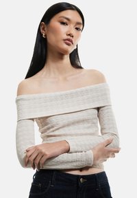 Beige off-the-shoulder toppi, jossa on pitkät hihat, kapea siluetti ja hienovarainen ribbaus, yhdistettynä tummiin farkkuihin.