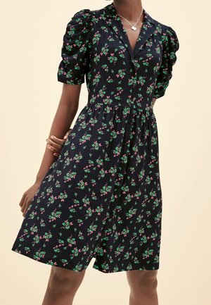 Sort kjole med pink og grøn blomsterprint, puffede korte ærmer, knapper foran og krave, båret med en sølv sommerfugle-halskæde.