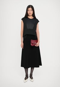 Melnā bezpiedurkņu blūze ar satīna priekšējo paneli, saskaņota ar melnu midi svārku un spīdīgu sarkanu rokassomu. Modelis valkā melnas zeķbikses un baltas kurpes.