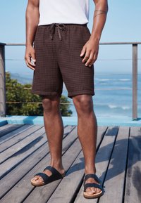 Brązowe szorty o fakturze, z elastycznym pasem i sznurkiem do wiązania, zestawione z czarnymi sandałami z korkową podeszwą, na drewnianym tarasie.