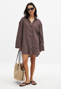 Abito camicia in lino marrone con bottoni frontali e maniche lunghe. Abbinato a una borsa a tracolla intrecciata bianca e nera e infradito.