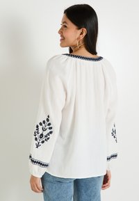 Blouse blanche avec broderie florale marine sur les manches et l'encolure, offrant une coupe décontractée et des détails froncés aux épaules.