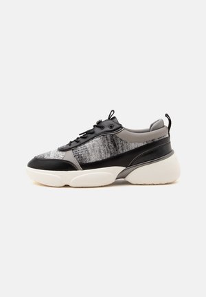 Zapatilla baja con paneles en negro, gris y plateado metálico, suela blanca gruesa, cordones y lengüeta en el talón, vista lateral sobre fondo blanco.