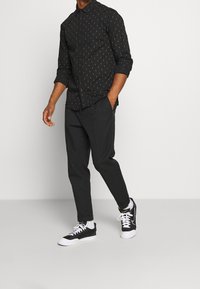 Camisa negra de botones con detalles en dorado, pantalones negros ajustados y zapatillas negras. La camisa presenta un patrón sutil y mangas enrolladas.
