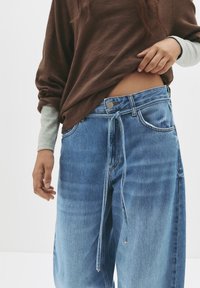 Blaue Denim-Hosenträgerjeans mit weitem Bein, Kordelzugbund und genähten Details. Kombiniert mit einem braunen Langarm-Oberteil mit grauen Ärmeln.