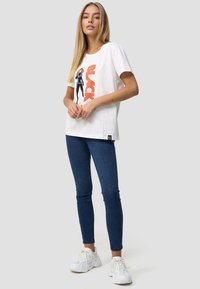 Hvid T-shirt med grafisk design i orange, parret med blå skinny jeans og hvide sneakers. Casual, afslappet pasform.