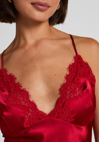 Κόκκινο σατέν καμαζόλ με λεπτή μαύρη δαντέλα κατά μήκος της γραμμής του λαιμού και απαλή, ελαφρώς γυαλιστερή υφή. Λεπτές ρυθμιζόμενες τιράντες.