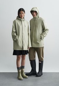 Πράσινες αδιάβροχες ζακέτες, γκρι παντελόνια και λάσπες. Οι μοντέλες φορούν χαλαρή εφαρμογή, με ρυθμιζόμενες κουκούλες και κούμπωμα με κουμπιά.