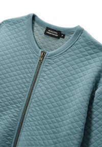Veste matelassée bleu clair avec un col rond, fermeture éclair à l'avant et tissu texturé présentant un motif en diamant. Étiquette visible avec la mention "MONOPRIX."