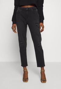 Jeans de mezclilla negra con detalles desgastados, corte recto, cinco bolsillos y cierre de botón, combinados con sandalias doradas con tiras.