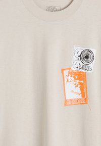 Beige Baumwoll-T-Shirt mit grafischen Drucken in Schwarz und Orange. Das Design umfasst ein Kamera- und Steckdosenmotiv mit einer strukturierten Oberfläche.