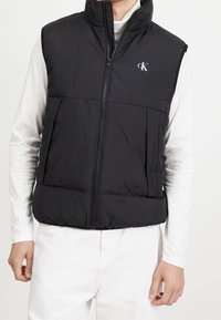 Schwarze Steppweste mit hohem Kragen und Reißverschluss, ausgestattet mit zwei Fronttaschen und einem dezenten strukturierten Muster. Logo auf der Brust.