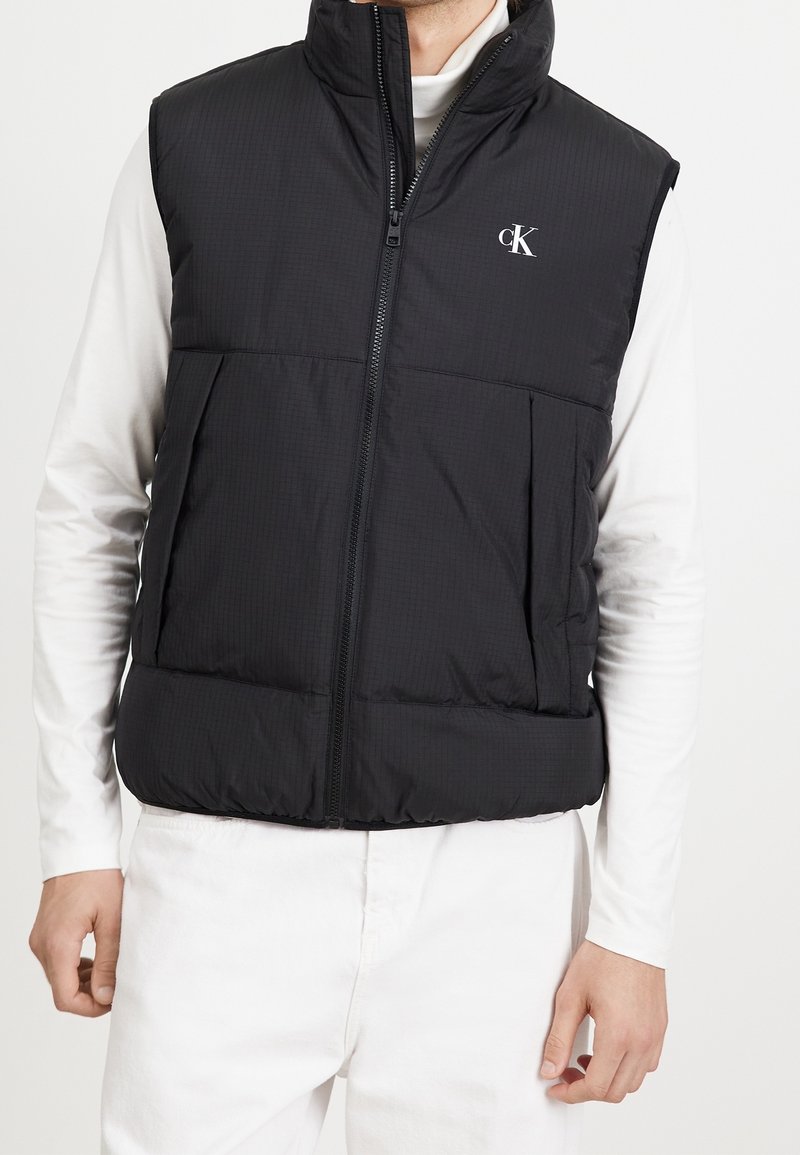 Schwarze Steppweste mit hohem Kragen und Reißverschluss, ausgestattet mit zwei Fronttaschen und einem dezenten strukturierten Muster. Logo auf der Brust.