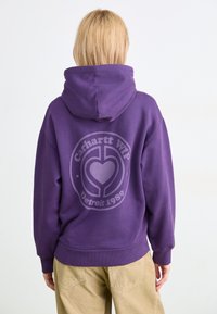 Violetti huppari, joka on valmistettu puuvillasekoitteesta. Hupparin takana on pyöreä logo painatus, jossa lukee "Carhartt WIP" ja "Detroit 1989" vaaleanharmaalla.