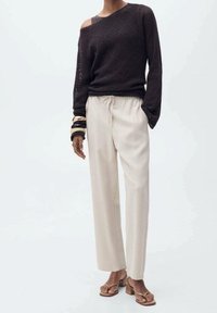 Donker gebreid trui met een off-shoulder ontwerp, gecombineerd met losse beige broek en tan sandalen; accessoires zijn onder andere gestapelde armbanden.