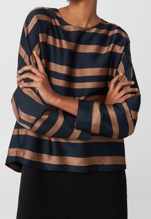 Personne portant un haut ample à manches longues rayé horizontalement bleu marine et marron, les bras croisés sur la poitrine.
