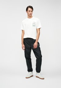 Λευκό βαμβακερό T-shirt με πράσινο κεντητό σχέδιο; μαύρο ελαφρύ παντελόνι σπορ; λευκά sneakers με καουτσούκ σόλες; μινιμαλιστική σπορ εμφανίσεις.