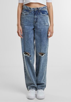 Personne portant un jean droit bleu clair déchiré aux genoux et des baskets blanches, debout devant un fond uni.