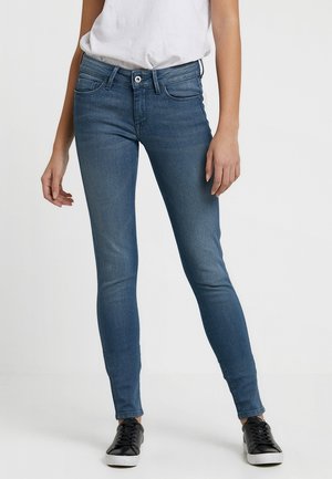 Vrouw draagt slim fit blauwe spijkerbroek, witte ingestopte t-shirt en zwarte lage sneakers tegen een effen lichte achtergrond.