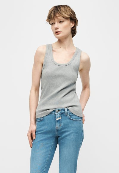 Jeune personne aux cheveux courts portant un débardeur gris sans manches et un jean bleu, debout avec une main dans la poche sur un fond uni.