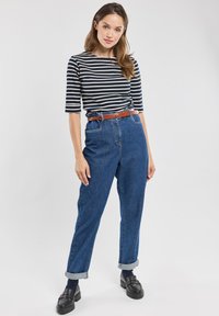 Camicia a righe blu e bianche a maniche lunghe, jeans blu a vita alta con risvolti, cintura in pelle marrone e calzature nere.