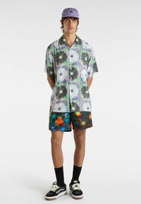 Camicia a maniche corte con un motivo floreale multicolore, abbinata a pantaloni corti multicolori e scarpe Vans nere, completata da un cappellino viola.