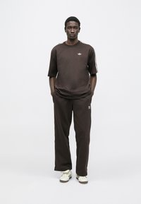 Barna rövid ujjú pamut póló kis fehér logóval a mellkason, hozzá illő barna nadrággal és fehér tornacipővel párosítva.