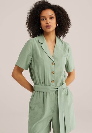 Ženska s kodrastimi lasmi, oblečena v svetlo zelen kombinezon s kratkimi rokavi, pasom in gumbi, stoji ob preprostem svetlem ozadju.