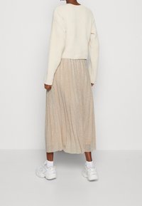 Frau in einem cremfarbenen Pullover, beiger, schimmernder, plissierter Midirock, weißen Socken und klobigen weißen Sneakern, die vor einem schlichten Hintergrund steht.