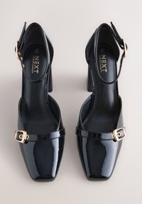 Black patent leather high heels with squared toes, ankle straps, and gold buckle accents. Smooth texture and glossy finish.