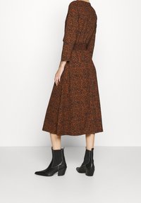 Frau in einem midi-langen, langärmligen Kleid mit braunem und schwarzem Muster, das eine taillierte Taille hat, kombiniert mit schwarzen, hohen Stiefeletten.