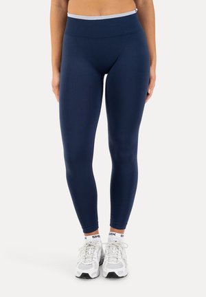 Kvinde iført marineblå højtaljede leggings, hvide mærkede ankelsokker og hvide sportssneakers, stående mod en ensfarvet baggrund.
