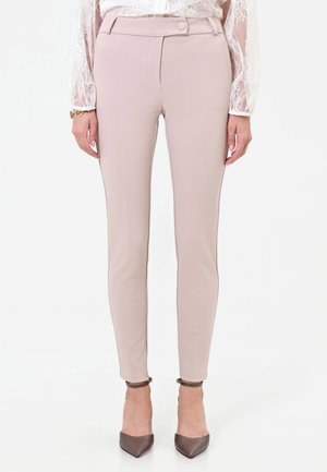 Donna che indossa pantaloni beige slim fit, camicetta bianca in pizzo, tacchi marroni con punta e cinturino alla caviglia, in piedi su sfondo bianco.