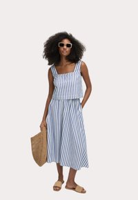 Gestreepte twee-delige outfit in blauw en wit, met een cropped top en een vloeiende rok. Accessoires bestaan uit een gevlochten tas en slip-on sandalen.