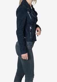 Navy suede biker jacket with silver hardware, zippered pockets, and a fitted design. Worn over gray jeans, showcasing textured material.