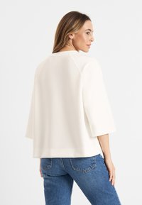 Crème-kleurige, losse top met driekwartmouwen en een lichte afgeronde onderkant. De stof heeft een gladde textuur, minimale naden en een ontspannen vorm.