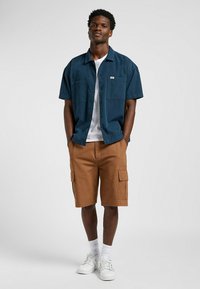 Camisa de manga corta azul marino con dos bolsillos frontales, combinada con pantalones cortos de carga beige y zapatillas blancas, con un ajuste casual y tejido texturizado.