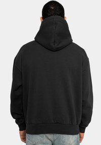 Svart hettegenser laget av mykt materiale med stor hette, senkede skuldre og ribbestrikkede mansjetter, båret over lyseblå jeans.