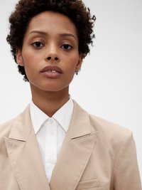 Blazer beige ajusté avec une finition texturée, revers crantés et poche poitrine simple, porté sur une chemise blanche à col avec fermeture à boutons.