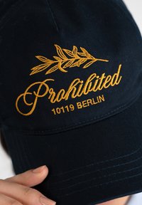 Tummansininen baseball-hattu, jossa on keltainen kirjailtu teksti "Kielletty 10119 BERLIINI" ja koristeellinen lehtikuvio.