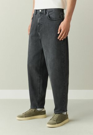 Man wearing loose-fit black denim jeans, white socks, and olive-green suede sneakers standing indoors against a plain background.