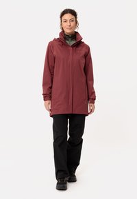 Giacca impermeabile bordeaux con cappuccio, dotata di zip anteriore, tasche laterali e polsini elasticizzati. Abbinata a pantaloni neri e stivali.